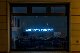 A window, looking in to a darkened office. On the window is a lit, blue neon sign which reads, “What is your story?”