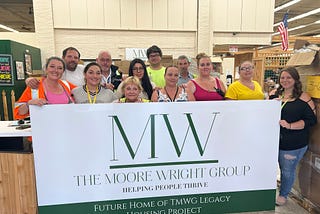 A dozen staff members from The Moore Wright Group pose together in the organization’s warehouse behind a white sign with green font announcing their Legacy Housing.