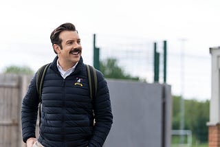 Ted Lasso (played by actor Jason Sudeikis) in a dark puffy coat wearing a backpack