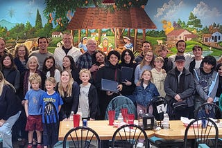 A large group of Youth Ambassador students and family members are gathered in front of table, posing for a photo. A mural is on the wall behind the group of a countryside scene.