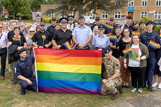 Group shot of MDP officers and staff standing with RAF Wyton personnel holding a rainbow flag, with buildings and trees in the background