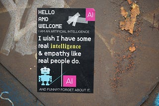 A sticker on a rusted metal background. The background of the sticker is black. The sticker shows a simple illustration of a small blue robot toy, the letters ‘AI’ in a pink square at the top and bottom, and the words, “Hello and welcome. Artificial intelligence. I wish I have some real intelligence and empathy like real people do. And funny? Forget about it.”