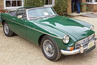 A British racing green MGB open-topped roadster sports car from the 1960s stands on gravel outside a house in England