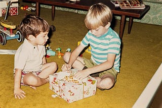 Two young boys sit on a mustard-yellow carpet, opening a wrapped gift together. One boy wears a light short-sleeved shirt and patterned shorts, while the other wears a green-and-white striped T-shirt with khaki shorts. A tricycle, toy ducks, and a wooden wagon are visible in the background.