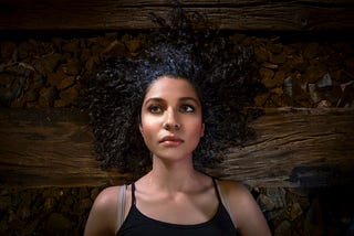 Woman of color lying on a wooden floor, black curly hair splayed around her head, gazing off into the distance, looking tired and contemplative