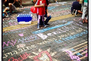 A young girl poses with a smile on a street filled with colorful chalk art and positive messages. She holds a red balloon and is surrounded by other participants engaged in creating chalk drawings. The vibrant artwork on the pavement includes uplifting words and drawings, creating a lively and creative atmosphere