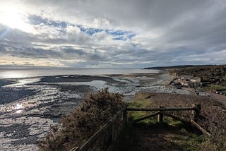 View from the cliffs at Llantwit Major beach at low tide, with rocky tidal flats, dramatic cloudy sky, sunlight on the sea, and a small building by the shore.
