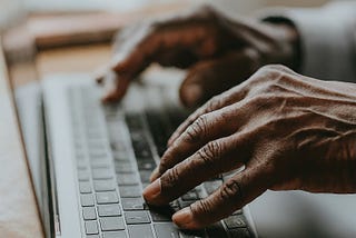 Close-up of African American senior’s hands typing on a laptop, with a softly blurred background.