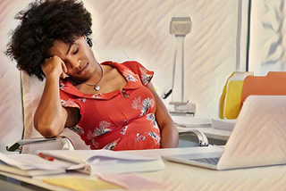 A creative writer appearing to be suffering from burnout and mental exhaustion sits behind a laptop. She is a black woman holding her head in her hand. She seems as if she isn’t inspired. The image represents lack of inspiration and productivity.
