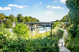 walkway alongside a pond that is surrounded by greenery with a lady and a little girl walking. Walkway leads to a train overpass in the background