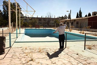 On a sunny day under a clear blue sky, an 80 year old man looks over an empty public swimming pool from behind a waist level chain link fence. The pool is large, painted bright blue and surrounding floor is made of flat polished stone. This man has not been here in almost 70 years. He is reminded of his childhood.