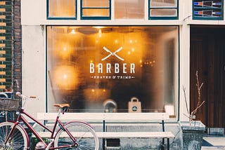 Outside of a barber shop in winter with warm lighting shining outward and a red vintage bicycle parked in front.
