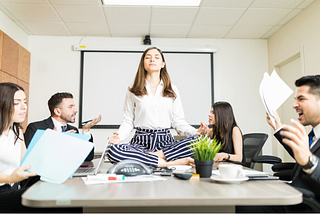 Woman sitting on conference room table in yoga pose while others in the meeting yell