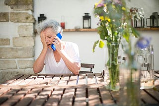 Old woman sitting at a table with one hand covering half her face and the other holding a bright blue phone to her ear. She looks distressed.
