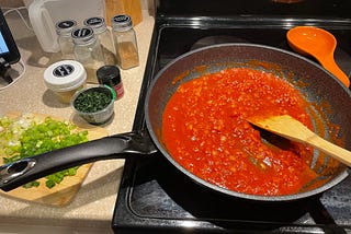 Pan on stove with red sauce and spatula in it and a collection of ingredients on the counter to the left