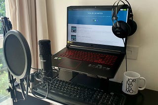 Foto da estação de trabalho, com uma mesa, um notebook suspenso por um suporte, um microfone preso por uma haste, um teclado, um fone de ouvido grande pendurado no notebook, e uma caneca branca escrito Jornalismo.