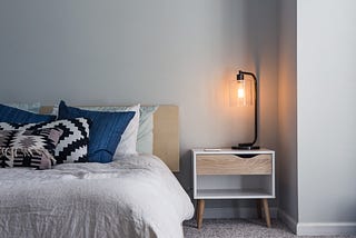 A low bed with a white bedspread and blue accent pillows. Next to it is a simple nightstand with a metal arm lamp with a clear shade, with a lit yellow light bulb inside it. Indirect sunlight comes from off screen to the right, lighting the entire scene.