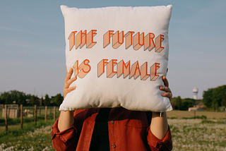 A woman holds a cushion with the words “the future is female” in front of her face. Photo by Sinitta Leunen on Unsplash