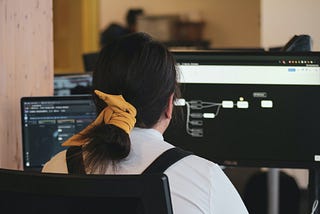 Woman with white long sleeve shirt with back turned and facing a monitor screen