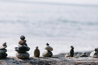 Stacks of balanced stones on a beach next to water. The picture is very calm and serene, in tones of light grey and subtle blue.