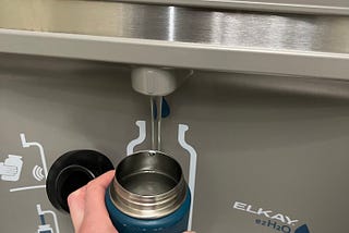 Photo of a hand holding a metal water bottle to refill at a water filling station at an airport. Alt text to demonstrate the importance of alt text.