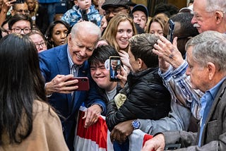 Joe Biden in a blue suit smiles, posing for a selfie with a supporter as a crowd surrounds him