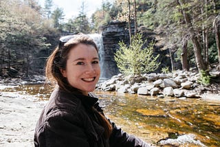In foreground, portrait of white woman’s head and torso. Long brown hair, big smile, pale skin. Solar shield shades on head. Background is a small waterfall in Hudson Valley New York. Rushing clear water, sunbleached rocks. A squat tree with fresh green leaves.