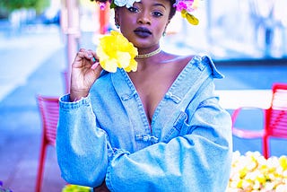 A woman dressed in denim stands with colorful flowers in her hair.