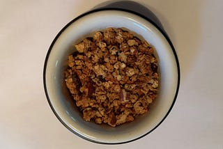 A small white bowl of granola sits on a small black plate with a white background.