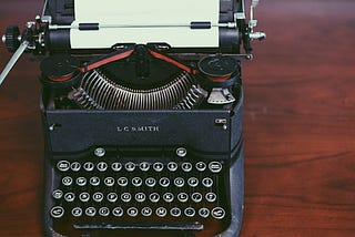 A cool black photo of a vintage typewriter sitting on a wooden desk. There’s a sheet of paper waiting to be filled with words.