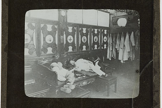 Black and white photograph of two men lying on a wooden bench inside a room. Slide is labelled ‘Opium Smokers’