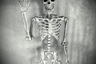 Grungy looking image of a human male skeleton with one arm raised like he is waving HI set against a black & white grainy backgound.