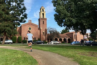 A runner turns a corner on a dirt track cutting through green lawn, in the shade of some trees