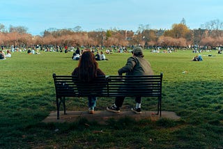 This picture shows a man and a woman sitting on a bench. We are looking at them from behind and we can see the park area they are facing that appears to have dozens of people enjoying nature. The man has his arm propped up on the back of the bench and his posture is turned towards the woman, indicating he may be talking to her even though he appears to be looking out away from her. She is facing straight, potentially not engaged, also looking out at the park.