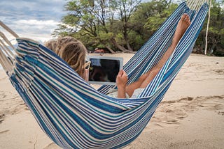 What No One Tells You About Remote Work and Being a Digital Nomad- A women laying in a hammock on the beach holding an ipad.