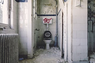 A dilapidated institutional bathroom with graffiti on the toilet and tank