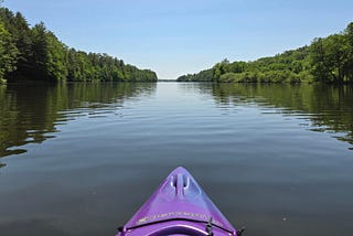 The nose of a purple kayak is centered with a wide lake channel ahead. Low hills covered in green forests are on both sides. The water is calm and there are no buildings, people, or other boats in sight.