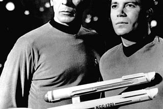 A black-and-white photo of Leonard Nimoy and William Shatner portraying Spock and Kirk. They are standing behind a model of the original USS Enterprise (NCC-1701).