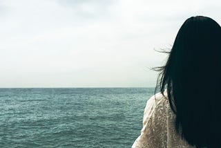 Photo from behind of a woman with long dark hair and a white cotton eyelet looking out at the horizon and the meeting of an aqua colored ocean and pale white skies.