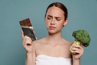 Young woman with acne holding a block of chocolate and broccoli