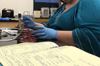 A woman in blue shirt wearing rubber gloves, working with Bunsen burner, while studying a book.