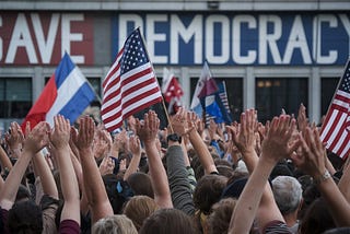 A photo of a large crowd of people with their hands raised in the air. There are multiple flags, including the American flag, being heldup by the crowd. The background contains a building with the words “Save Democracy” written in large letters.