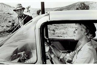 black & white image of a woman in the driver’s seat of a car with a male hitchhiker in the background on the passenger’s side