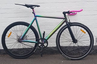 A fixed gear track bike in an obnoxiously flashy paint job braced against a while wall.