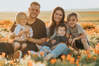 Family, 2 parents and 3 kids, sitting in a sunny field with lots of orange and white flowers