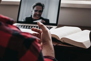 Two people are in a video meeting. The man on the laptop screen is wearing glasses and is smiling; the other person is gesturing with their back to the camera. There’s an open book next to the laptop.