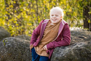 A boy with cancer posing outside by a rock.