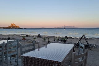 A taverna in Kefalos, Kos, looking out to sea. It’s sunset. There’s is a table in the foreground and islands on the horizon
