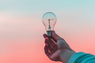 A hand is holding a light bulb in front of a gradiant that goes from a light, cool blue to a warm, fleshy pink.