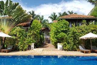 A tropical boutique hotel in Battambang, Cambodia, featuring a serene blue pool surrounded by lush greenery, palm trees, and traditional wooden villas with terracotta roofs, creating a tranquil and luxurious retreat.